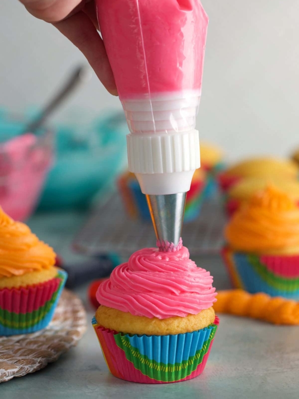 A piping bag is piping pink frosting onto a vanilla cupcake.