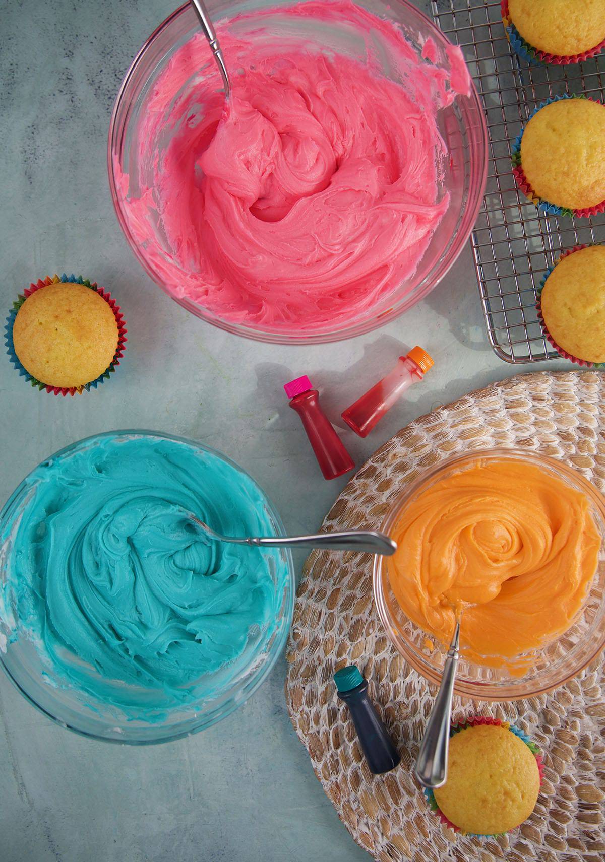 Three bowls contain blue, pink, and orange frosting.