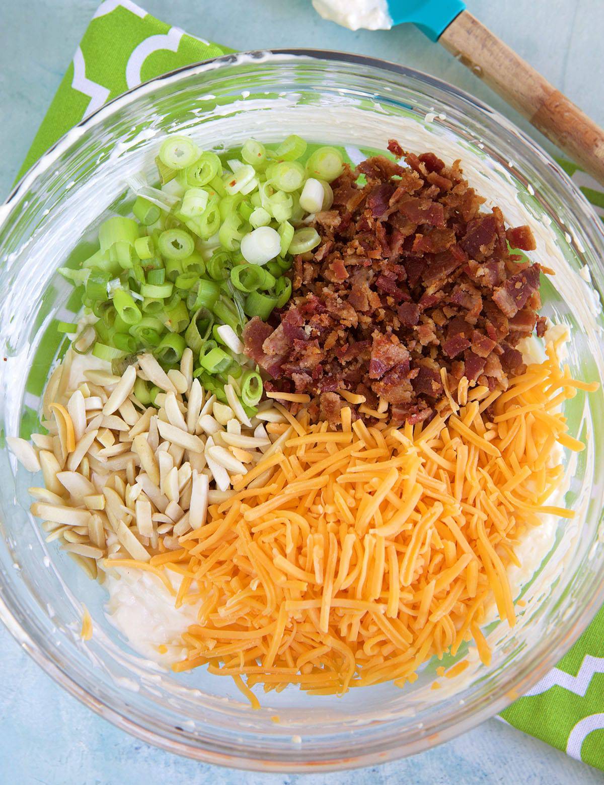 Almonds, cheese, bacon and green onions are placed on top of mayonnaise in a glass bowl.