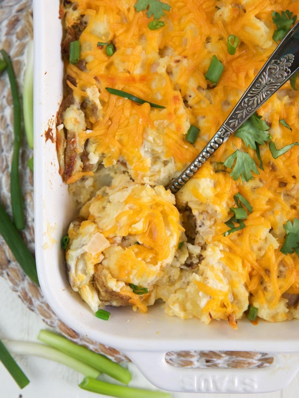 A large serving spoon is dipping into a casserole dish filled with cheesy potatoes.