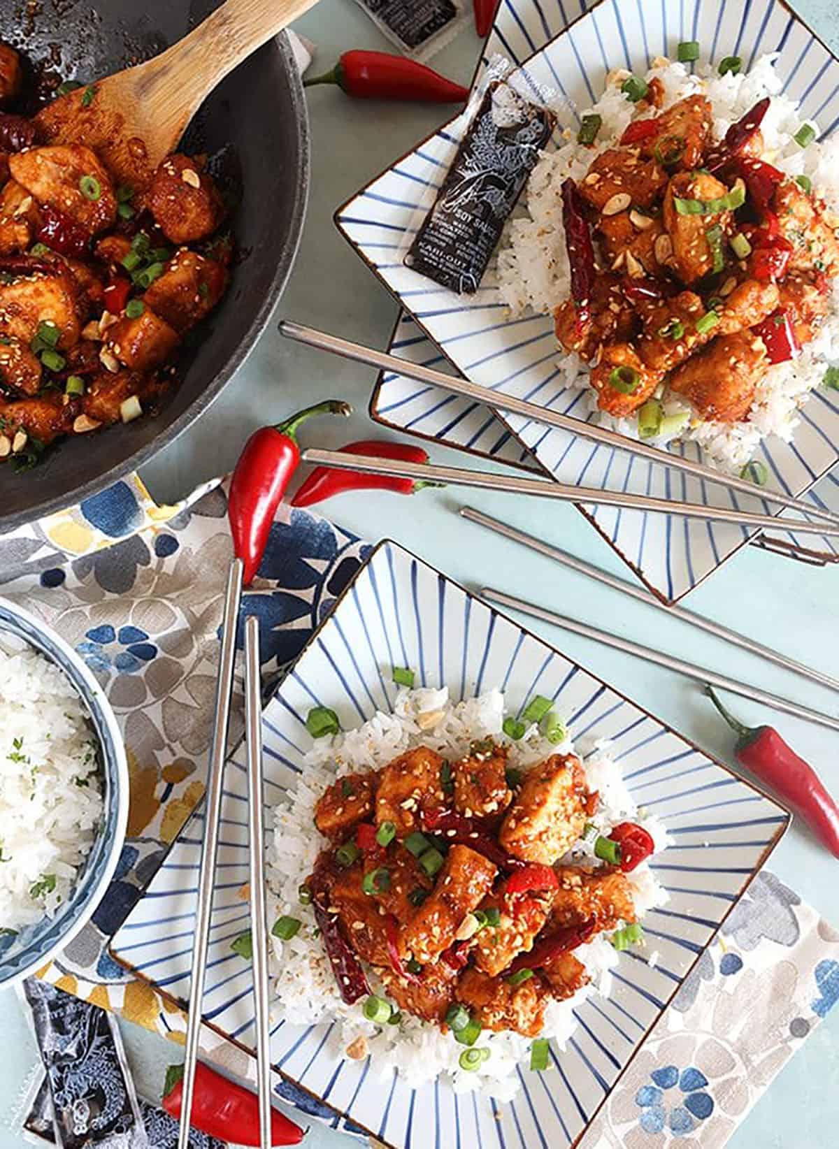 Overhead shot of szechuan chicken on two square plates with white rice and a wok.