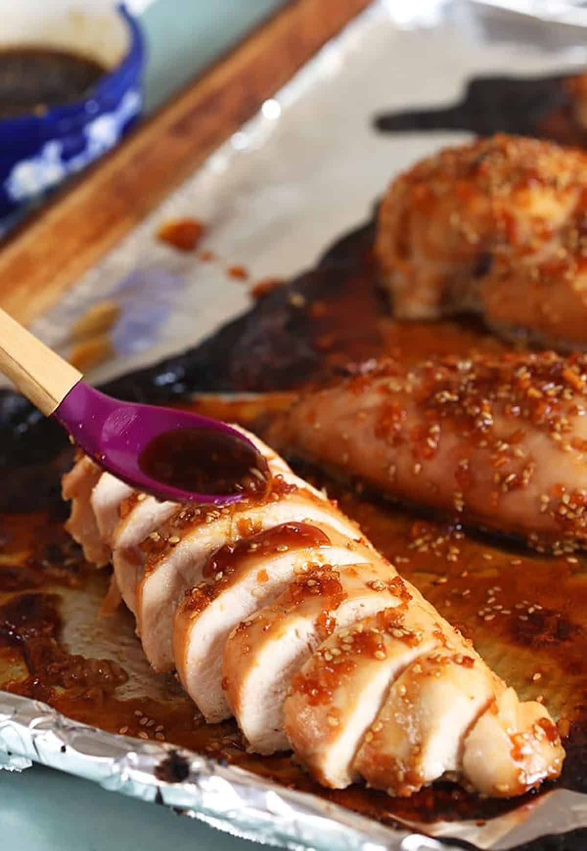 Sliced Teriyaki Chicken on a baking sheet with a spoon drizzling sauce over the chicken.