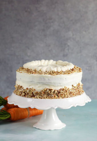 Whole carrot cake on a white cake pedestal on a gray background.