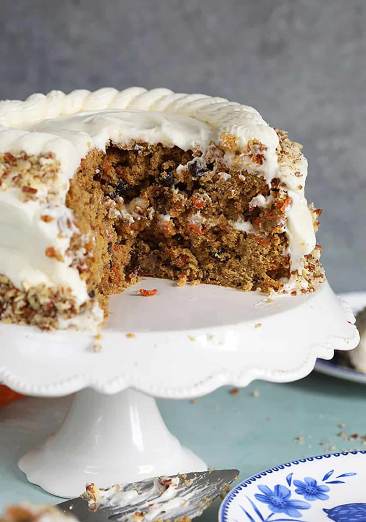 Carrot cake on a white cake plate with a slice taken out.