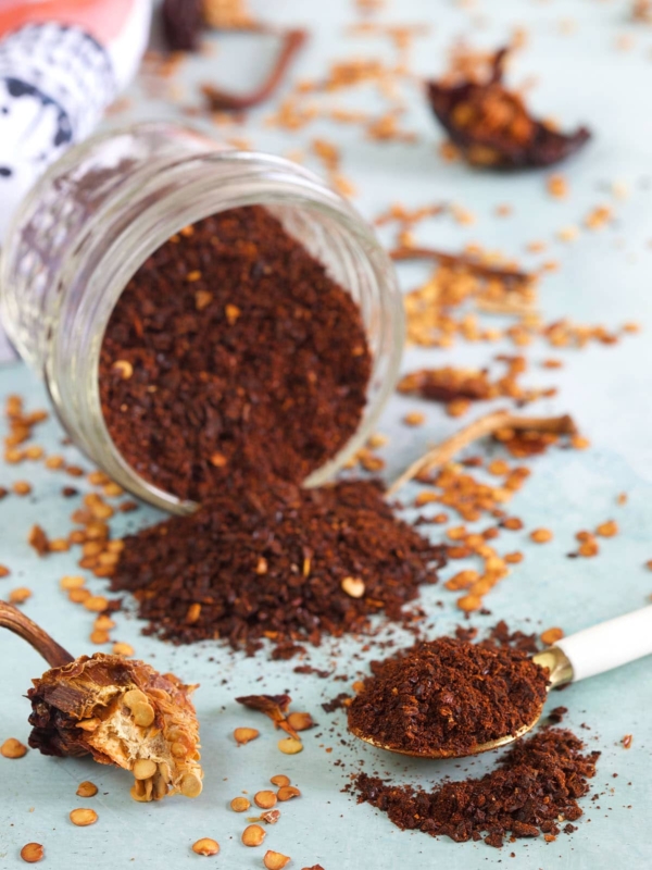 A spoon and jar both containing portions of chili powder are spread out on a white surface.