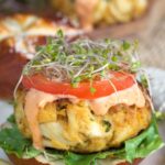 A crab cake is placed atop a bun and topped with tomato slice, sauce and micro greens.