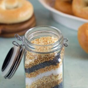 Everything Bagel Seasoning ingredients layered in a small jar on a blue background with mini bagels in the background.