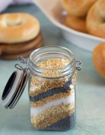 Everything Bagel Seasoning ingredients layered in a small jar on a blue background with mini bagels in the background.