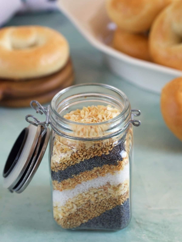 Everything Bagel Seasoning ingredients layered in a small jar on a blue background with mini bagels in the background.