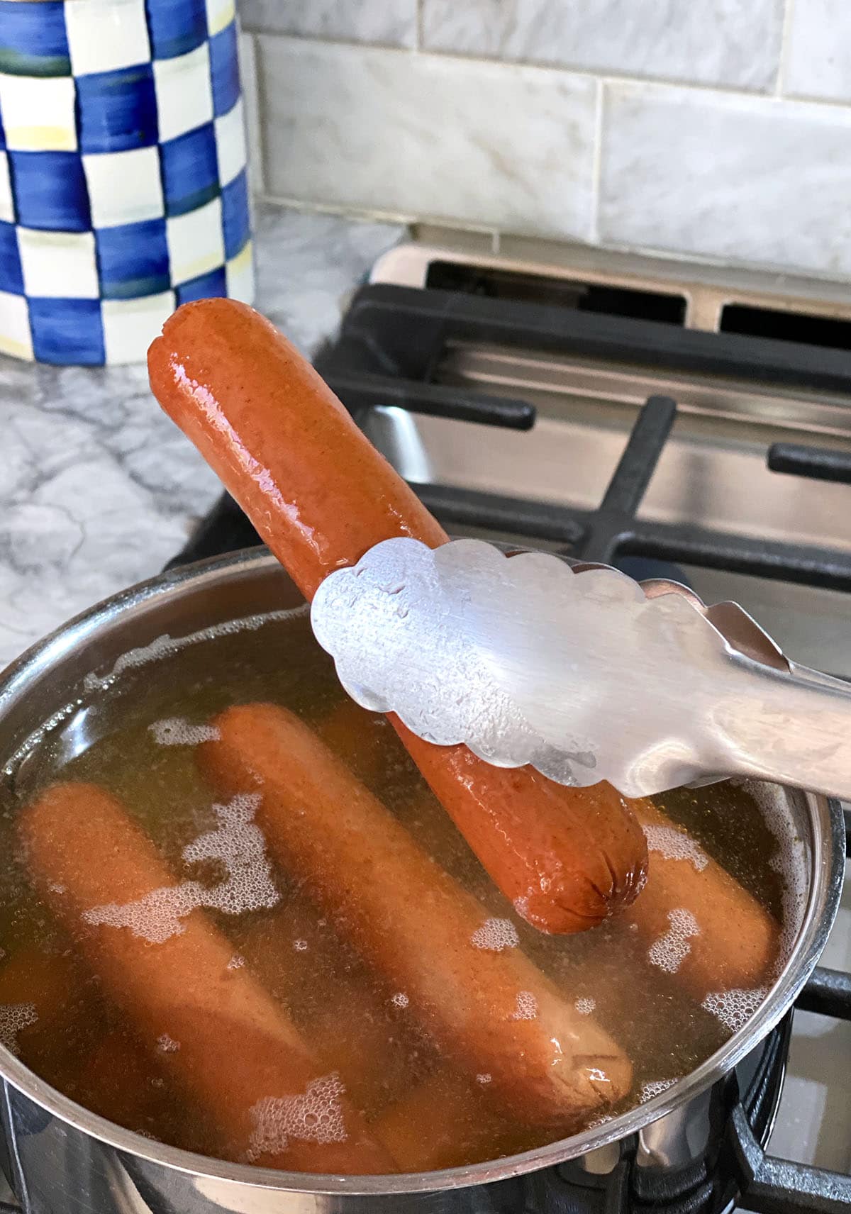 A pair of silver tongs is lifting a hot dog from a pot of boiling water.