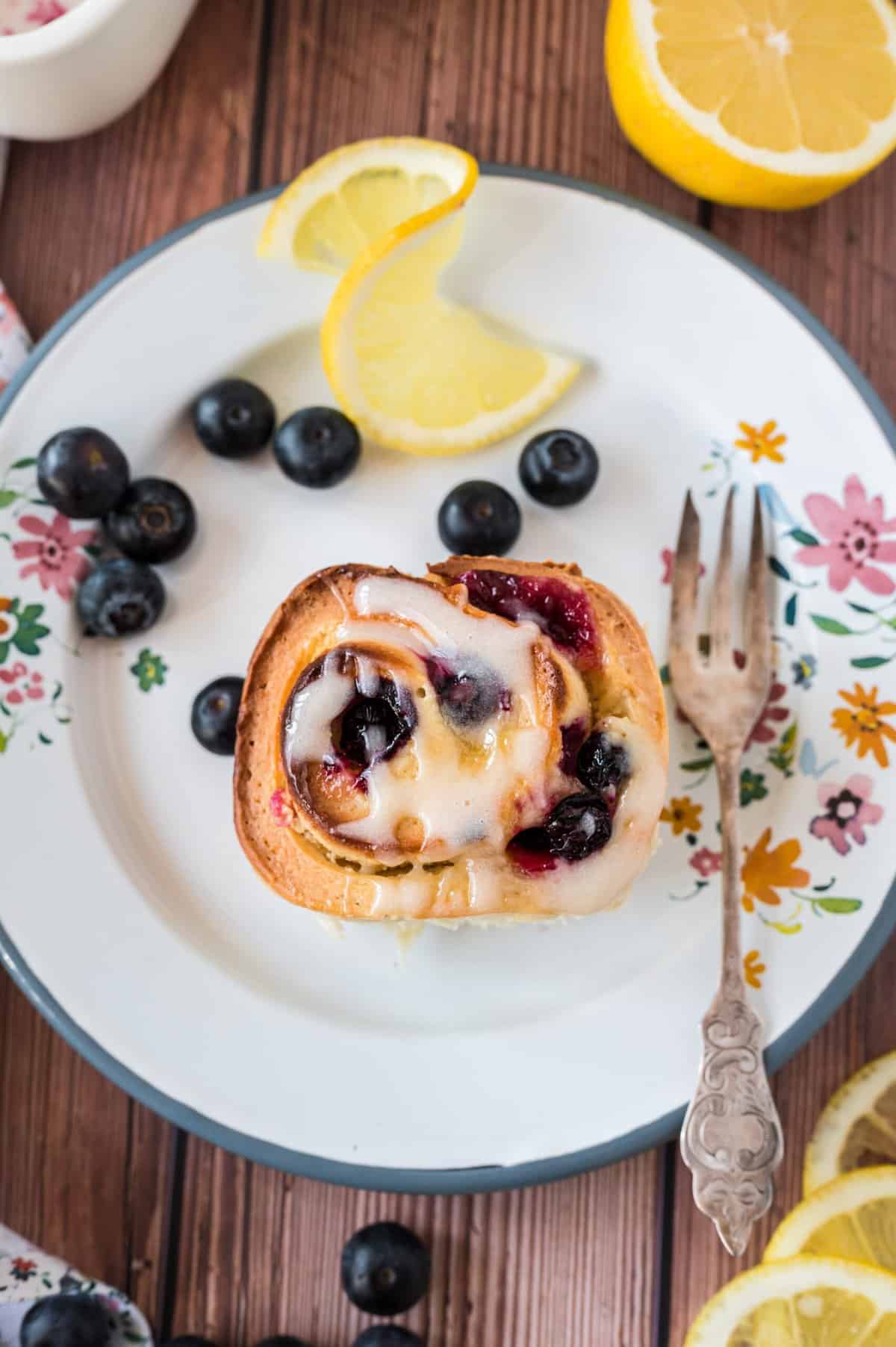 A lemon blueberry roll is on a white plate, next to a lemon slice and fresh berries.