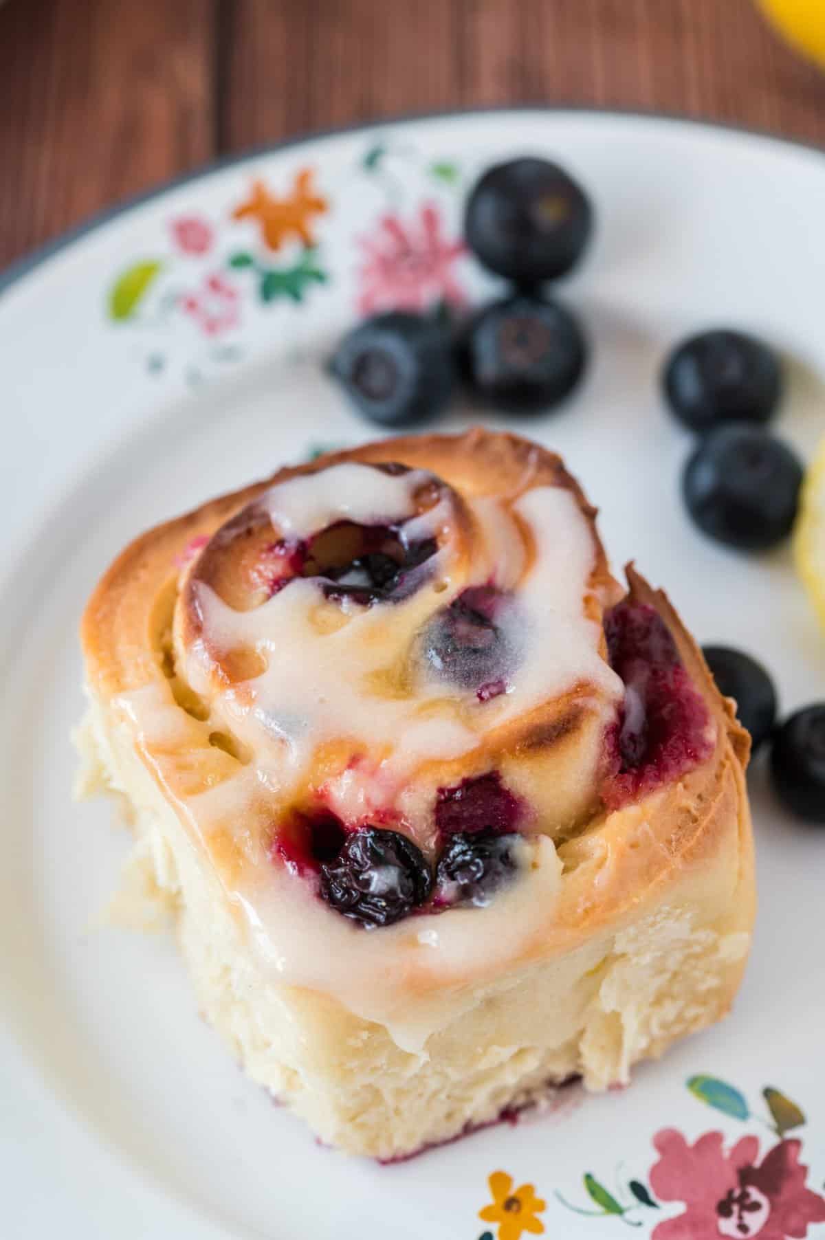 A glazed lemon blueberry roll is on a white plate with little decorative flowers.