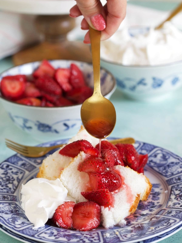 A spoon is drizzling macerated strawberries over a piece of cake.