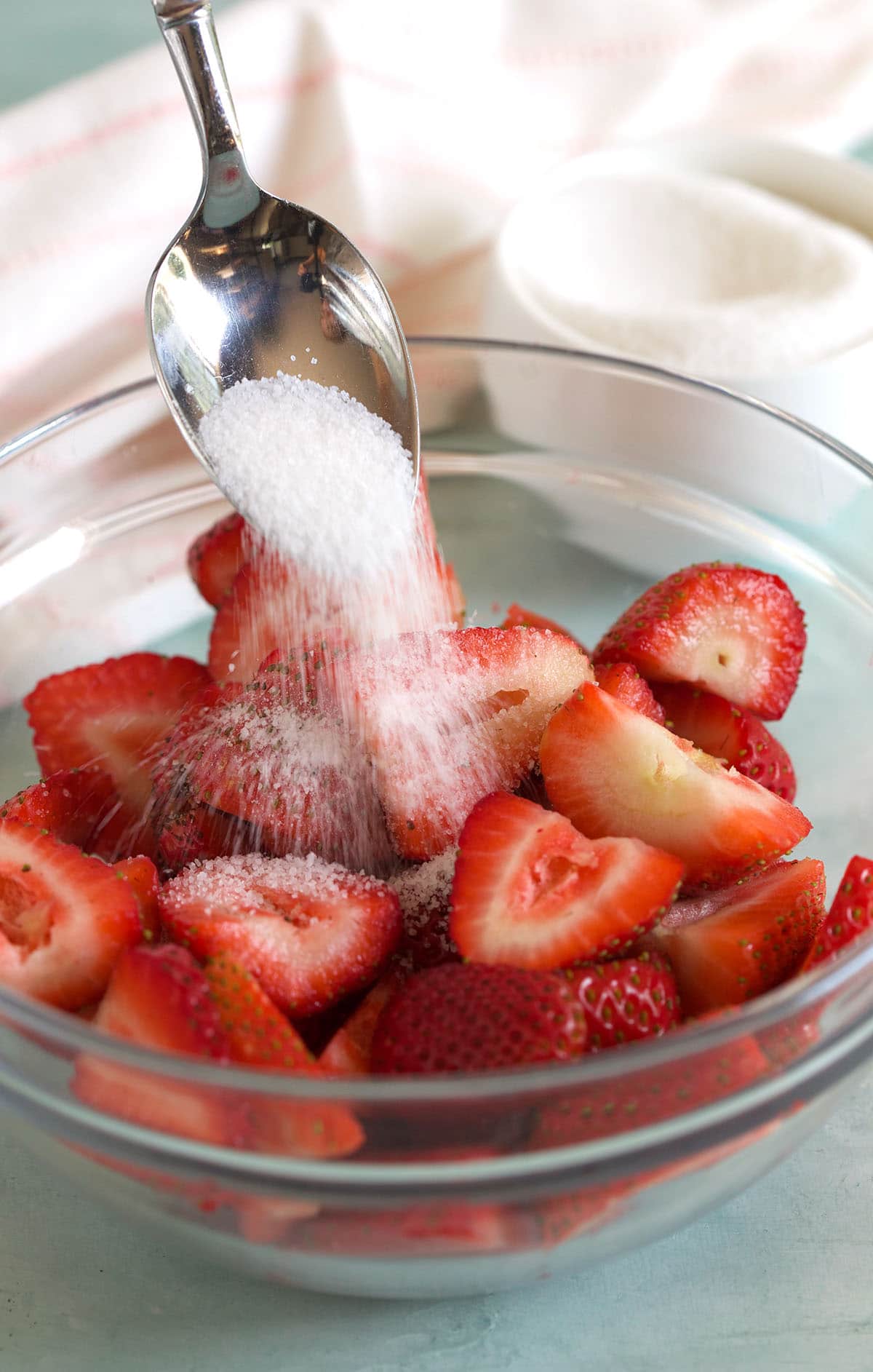 A spoonful of sugar is being poured on top of a bowl of fresh cut strawberries. 
