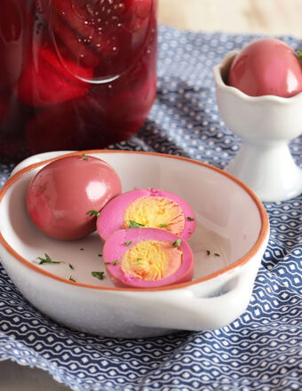 Beet Pickled Egg cut in half on a white dish with a whole egg behind it and a jar filled with pickled eggs in the background.