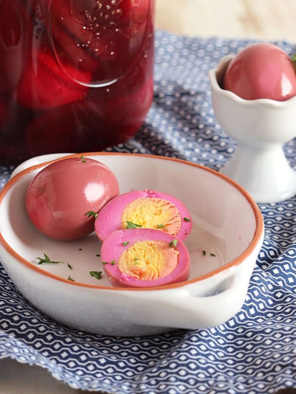 Beet Pickled Egg cut in half on a white dish with a whole egg behind it and a jar filled with pickled eggs in the background.