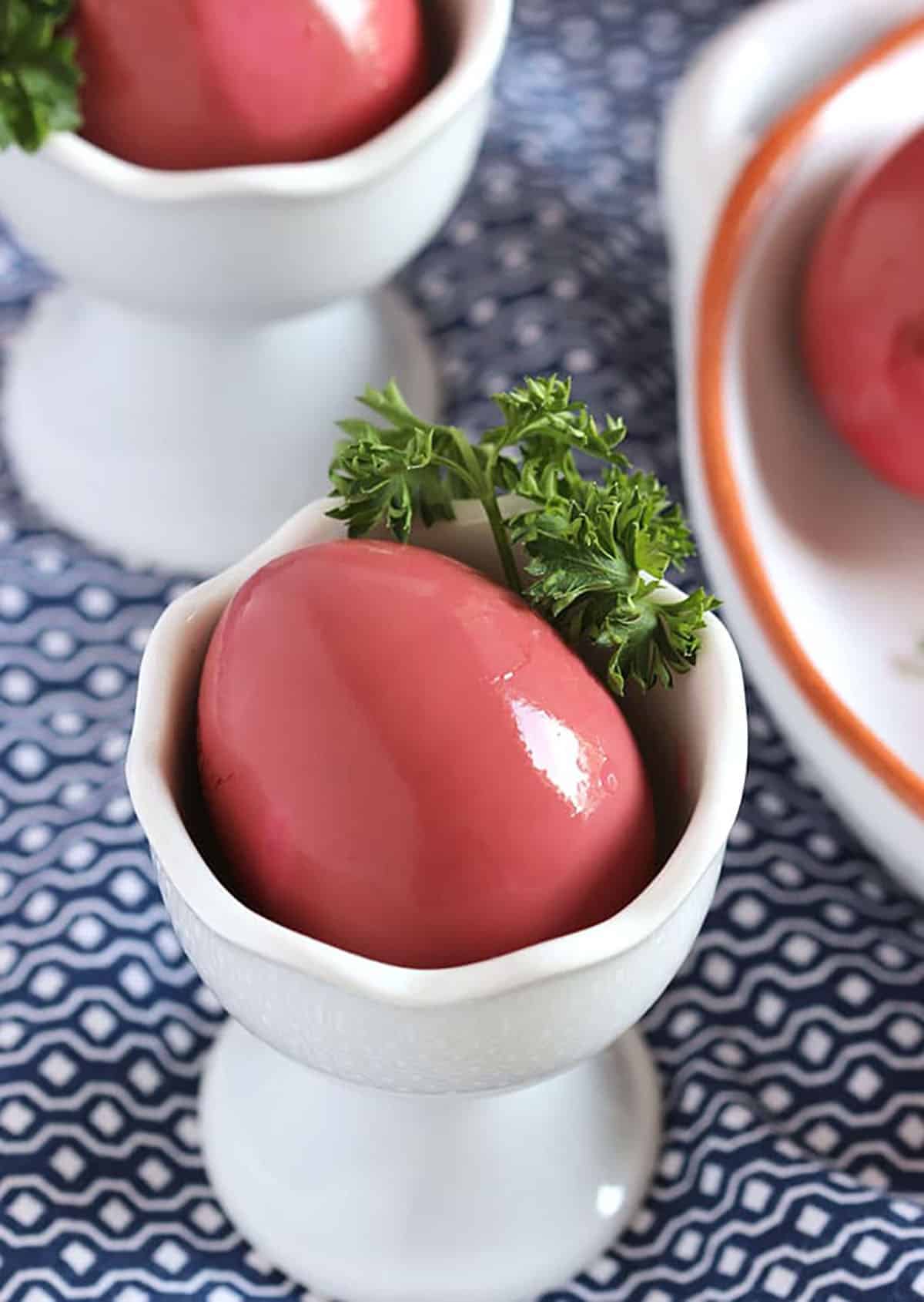 Beet pickled egg with a sprig of parsley in a white egg cup on a blue and white napkin.