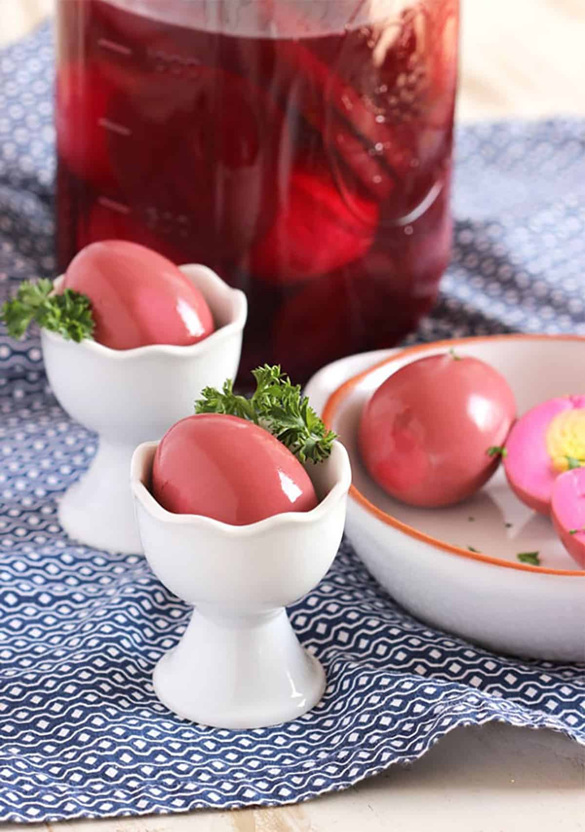 Two white egg cups with pickled beet eggs in them on a blue and white napkin with a jar in the background showing pickled eggs and beets in a jar.