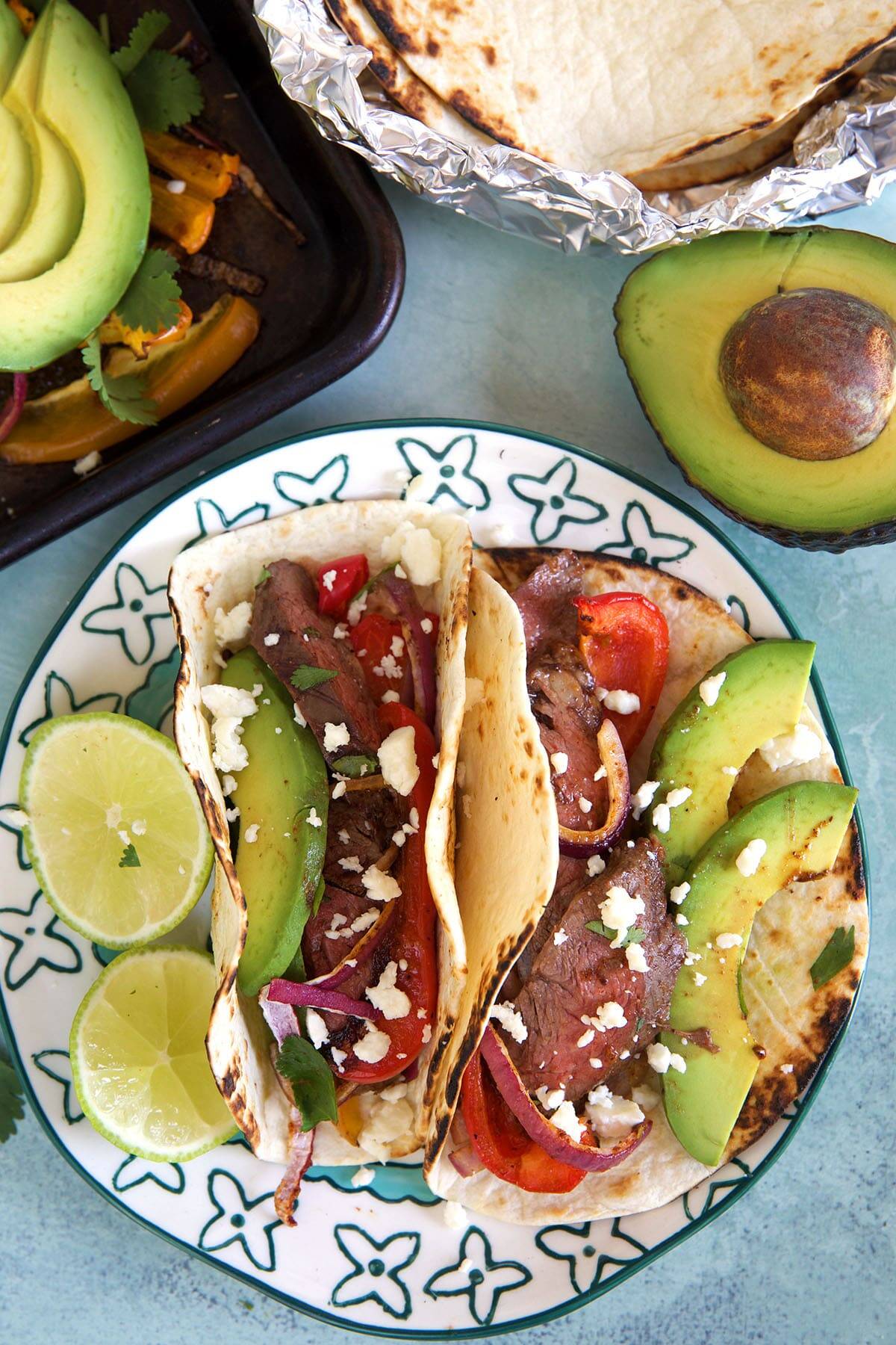 two steak fajitas on a plate on a white background with an avocado in the corner.