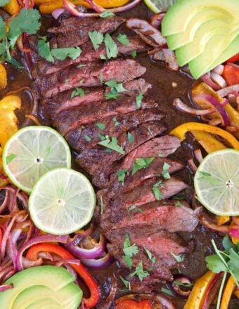 Sliced steak on a sheet pan with fajita vegetables, lime and avocado.