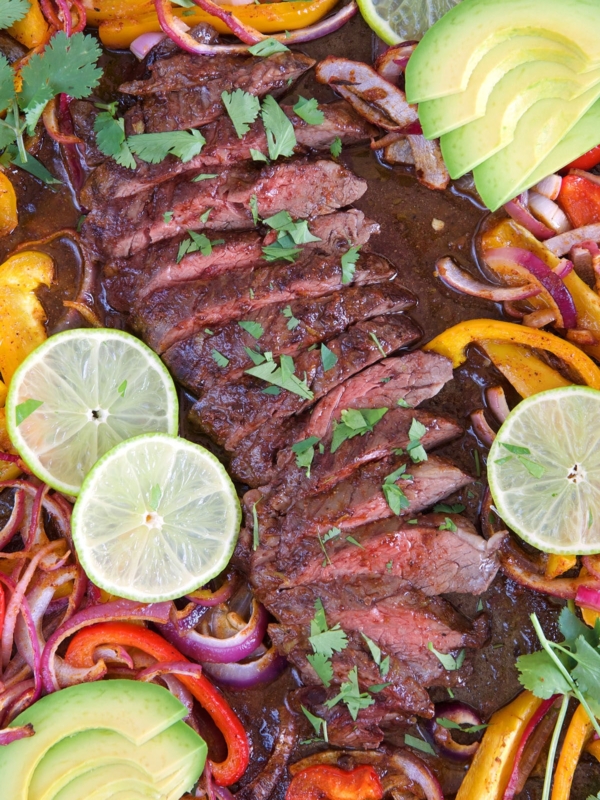 Sliced steak on a sheet pan with fajita vegetables, lime and avocado.