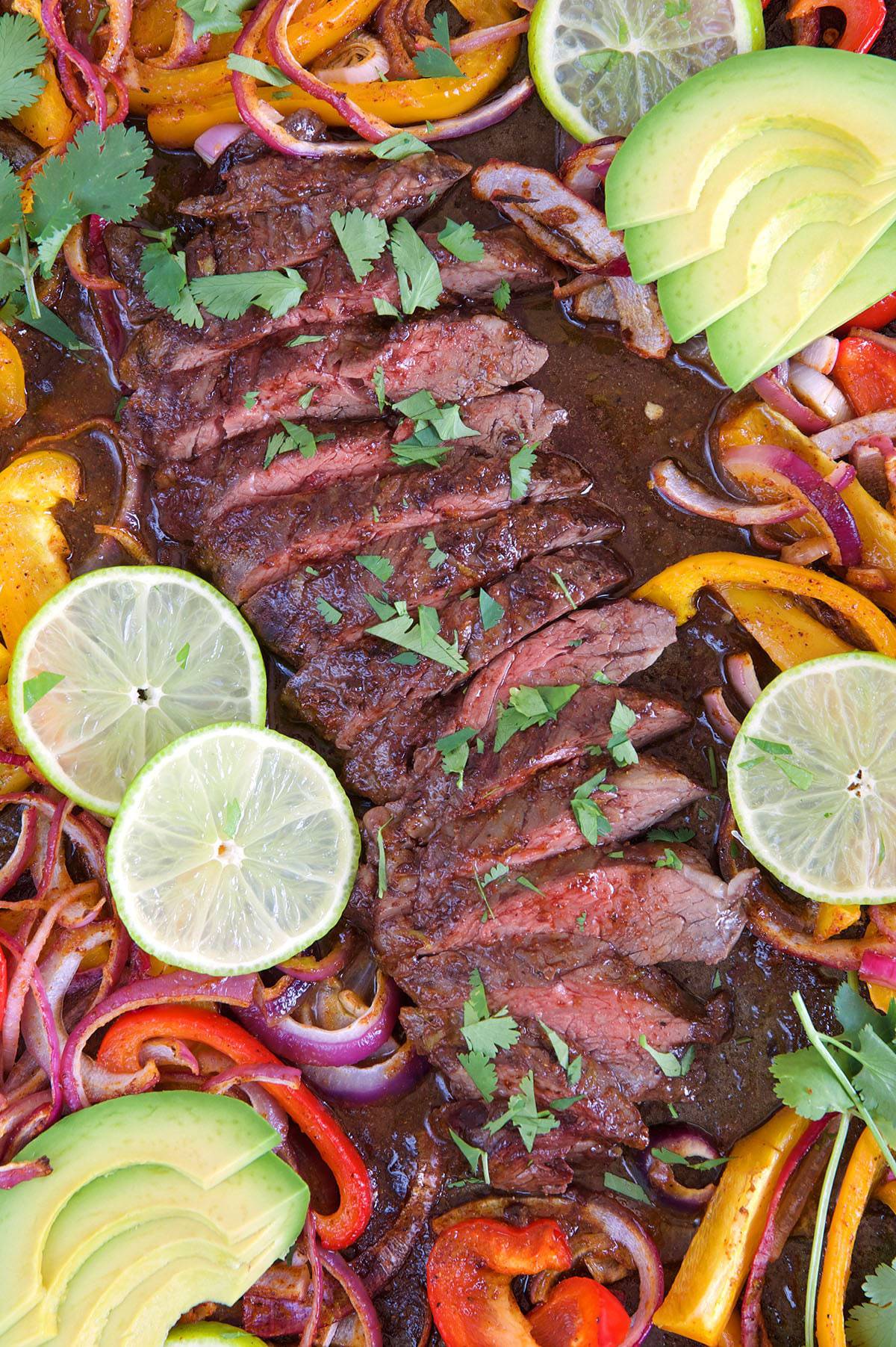 Sliced steak on a sheet pan with fajita vegetables, lime and avocado.