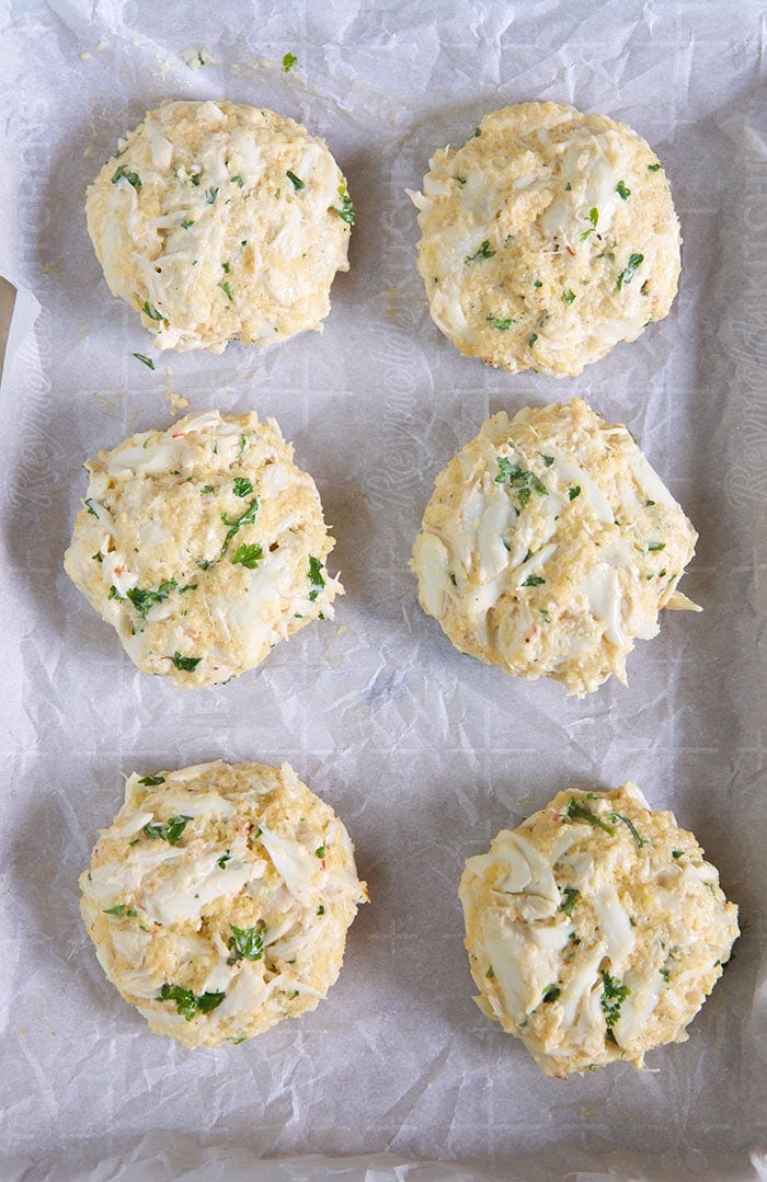 Six crab cakes are placed on a sheet of parchment paper.