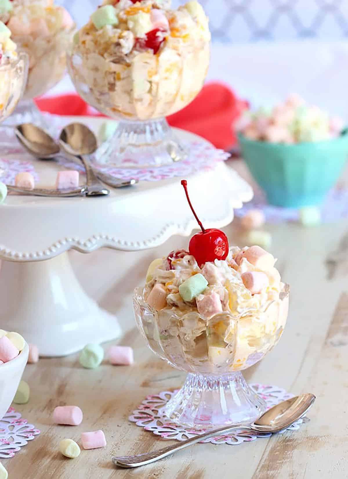 Ambrosia Salad in a glass dessert cup with a cake stand in the background with more bowls of ambrosia in them. 