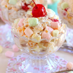 Ambrosia Salad in a glass bowl with a cherry on top and a spoon in front of the bowl.