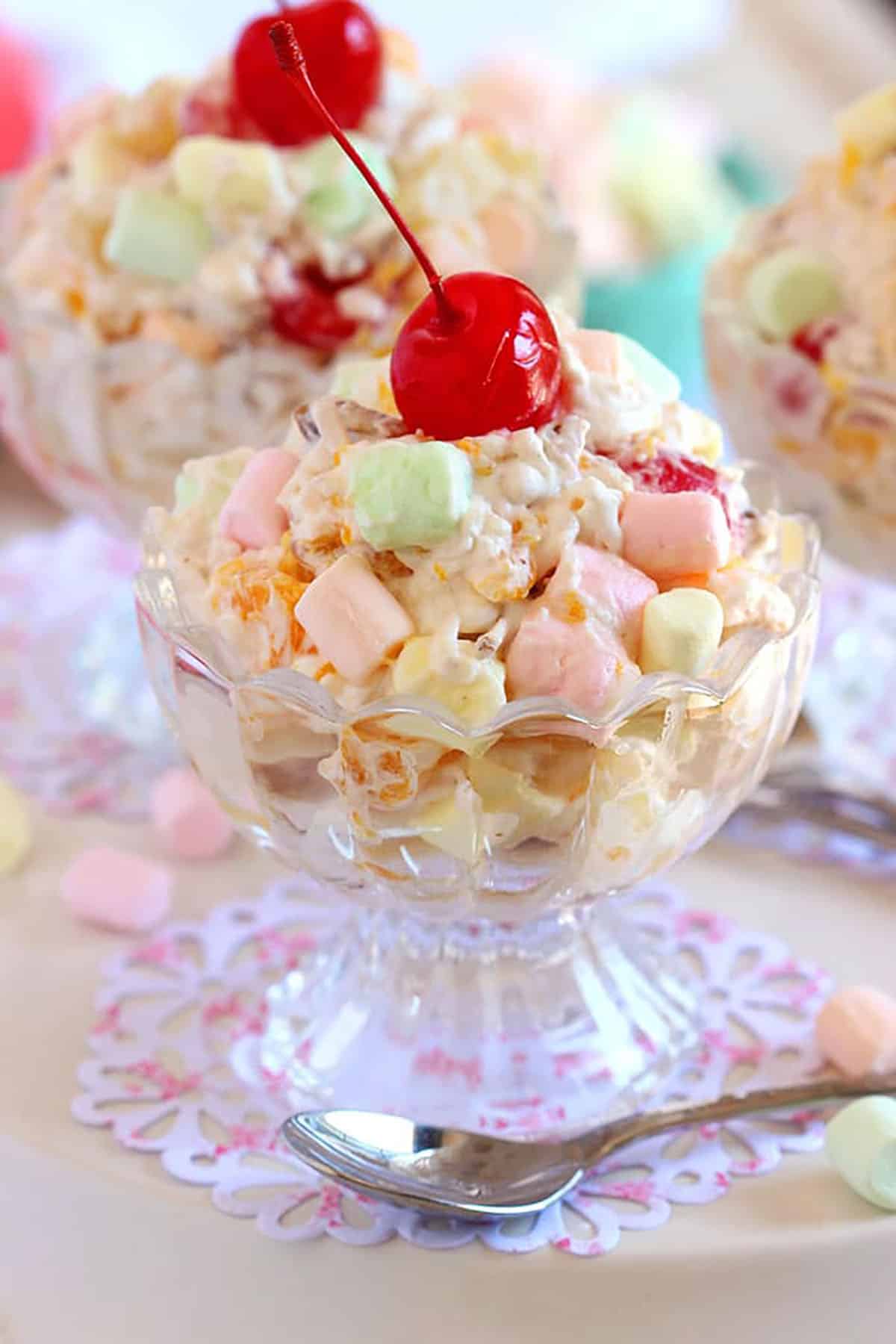 Ambrosia Salad in a glass bowl with a cherry on top and a spoon in front of the bowl.