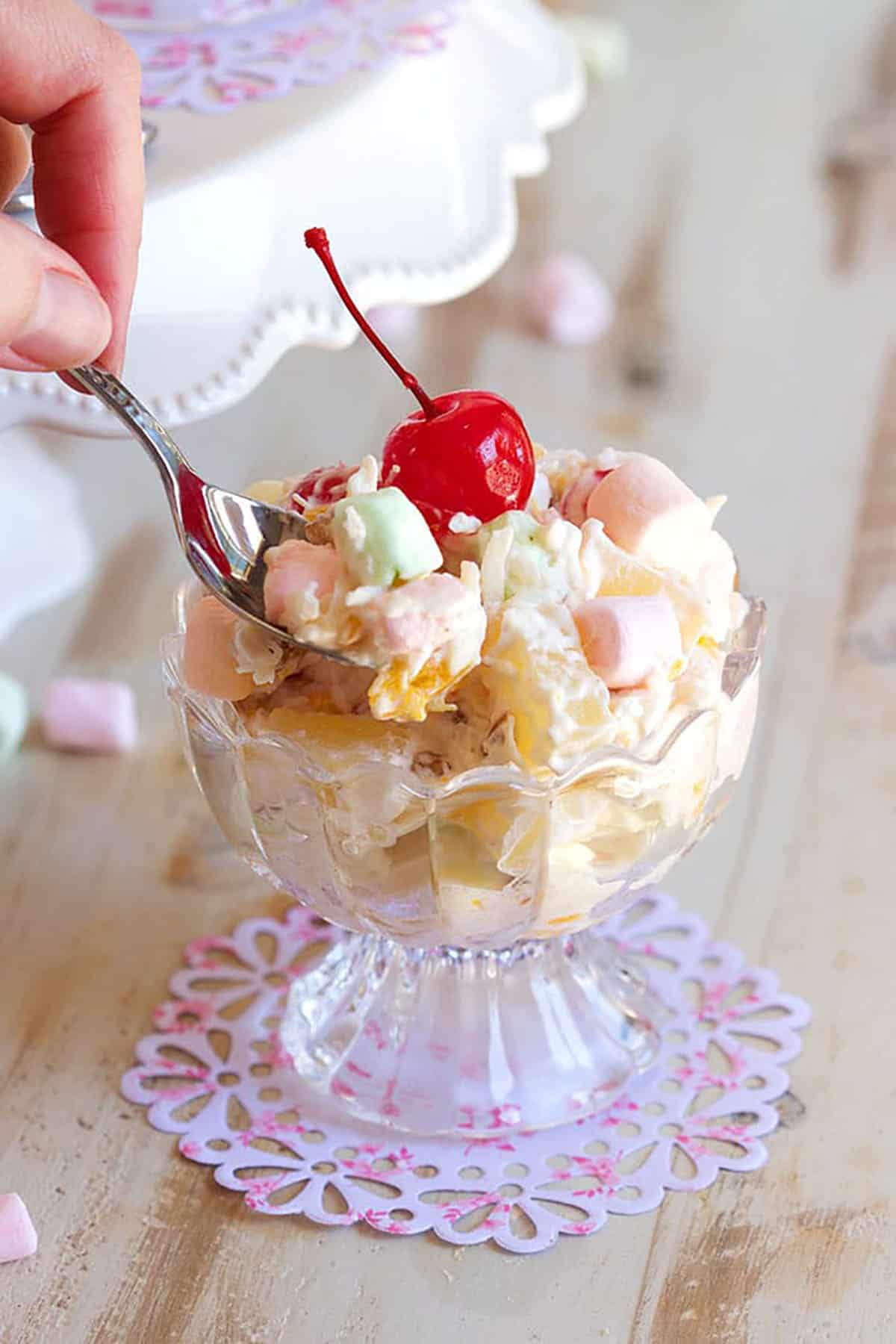 Glass dessert bowl with ambrosia salad topped with a cherry and a hand dipping a spoon into the salad.