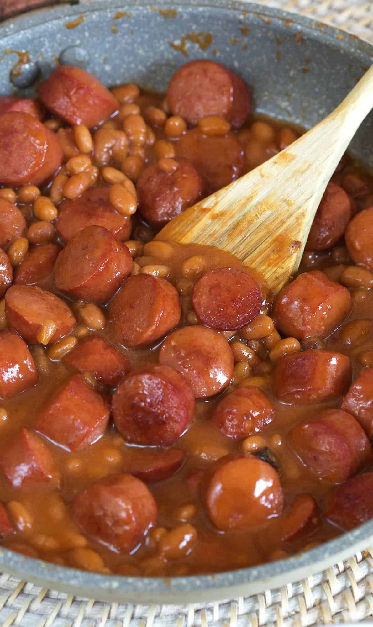 A wooden spoon is placed in the middle of a skillet filled with hot dogs and beans.