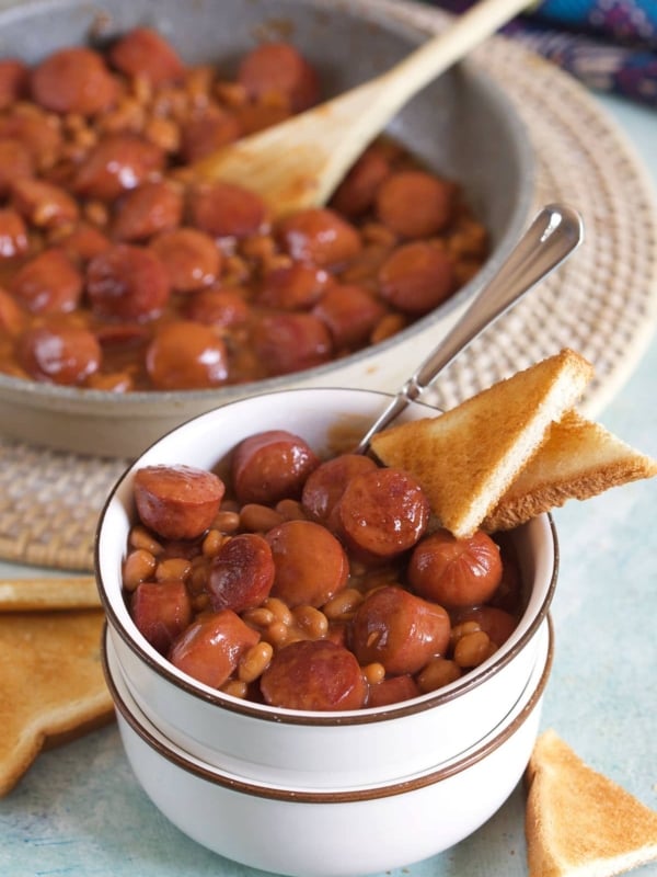 A small white bowl contains a portion of beanie weenies and two small pieces of toast.