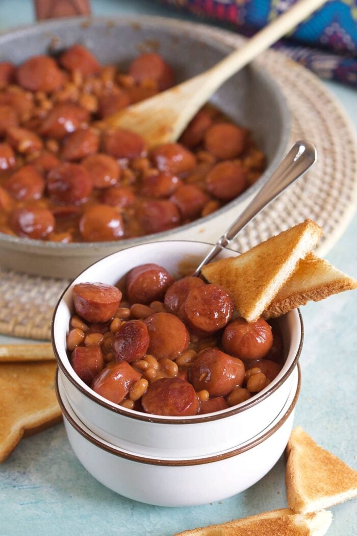 A small white bowl contains a portion of beanie weenies and two small pieces of toast.