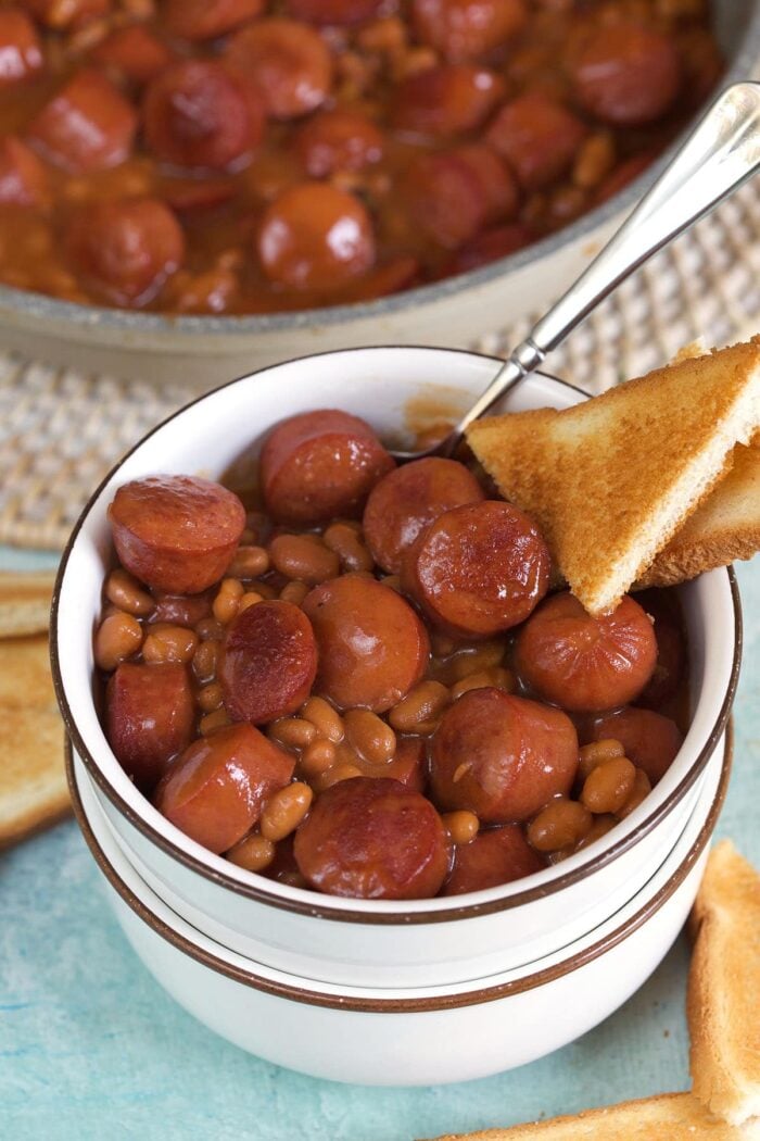A serving of beanie weenies is in a small white bowl with a slice of bread on the side.