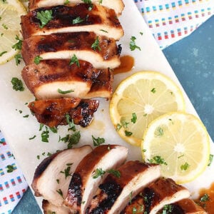 Two sliced marinated chicken breasts on a white marble platter with lemons