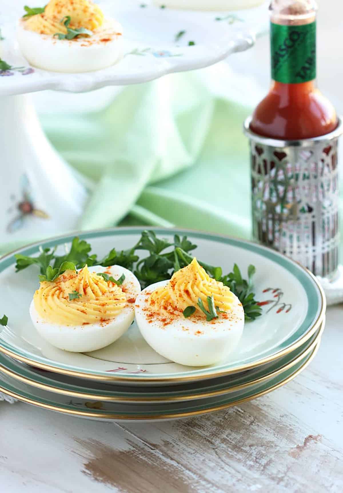 Two deviled eggs on a pile of plates with a bottle of hot sauce in a silver holder.