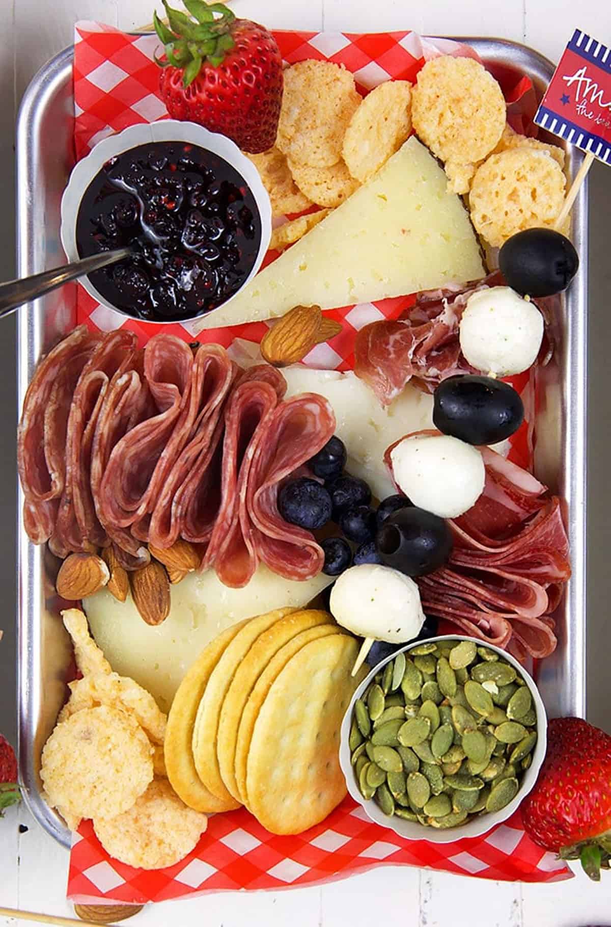 Overhead shot of charcuterie items on a stainless steel board with a red and white paper under the cheese.