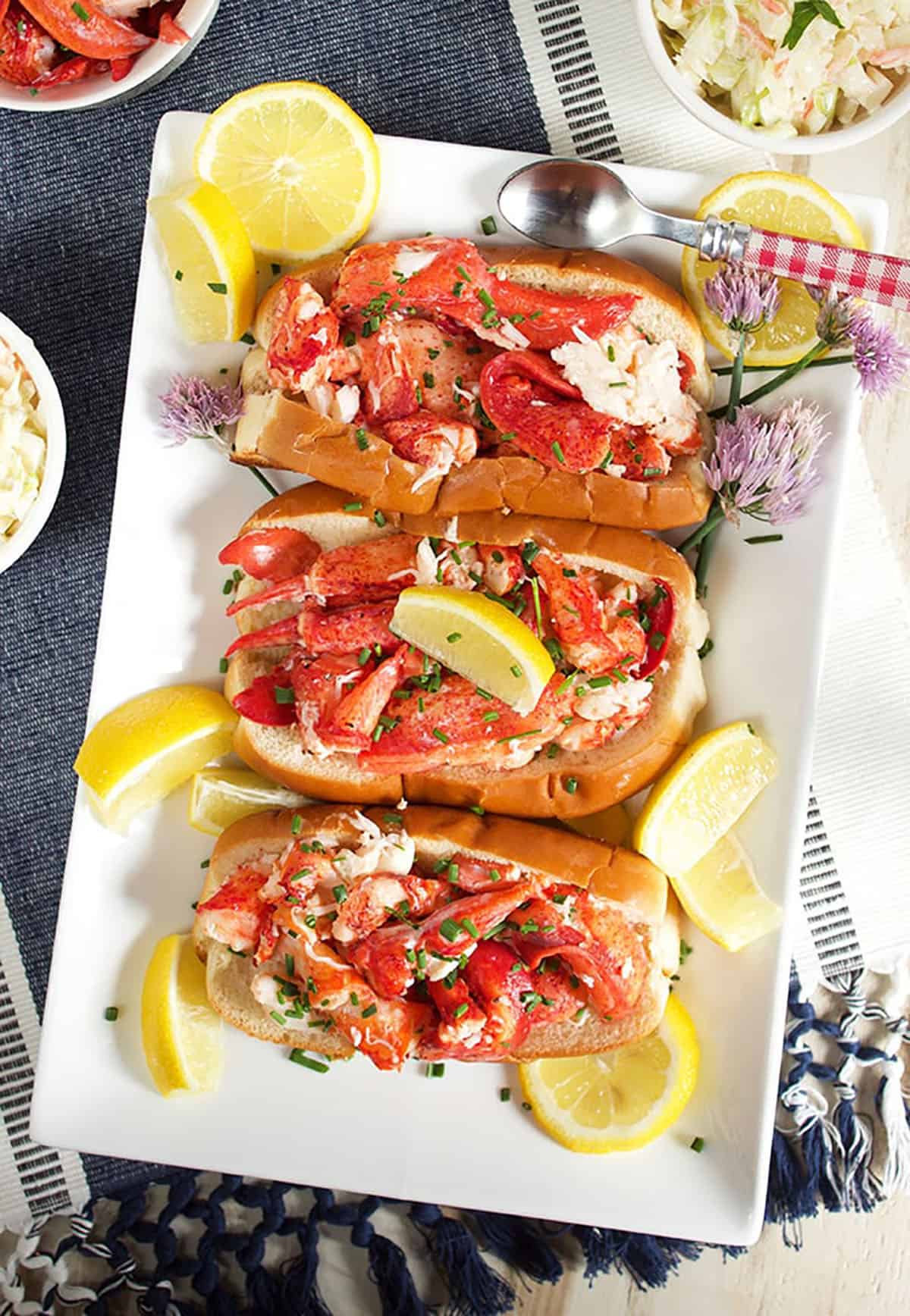 Three lobster rolls on a white platter with lemon wedges