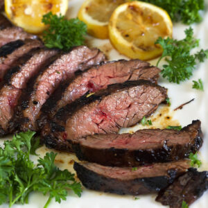 A sliced steak is placed next to herbs and lemon halves on a white plate.