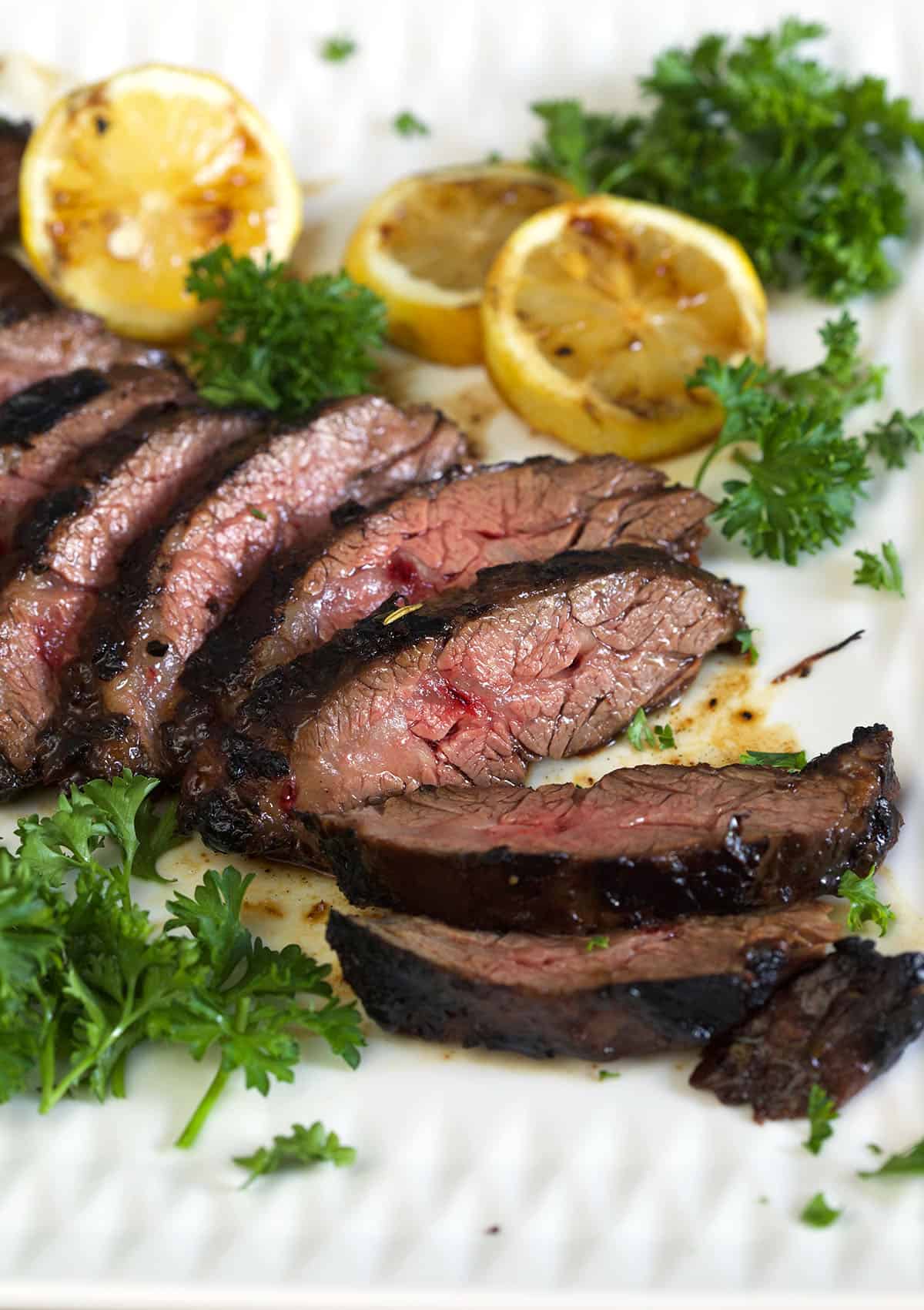 A sliced steak is placed next to herbs and lemon halves on a white plate. 