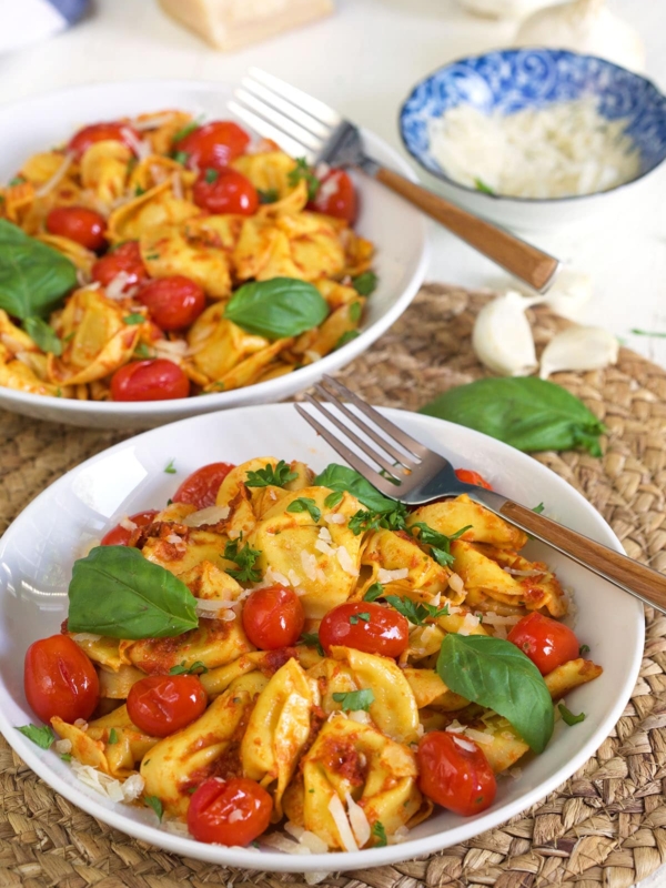 Two white bowls are filled with cheese tortellini and tomatoes.