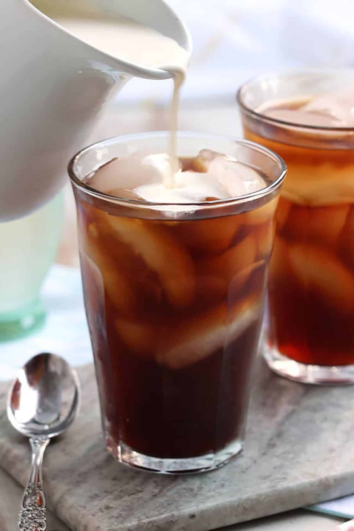 cream being poured into a glass of cold brew coffee with ice and a spoon to the left of the glass.