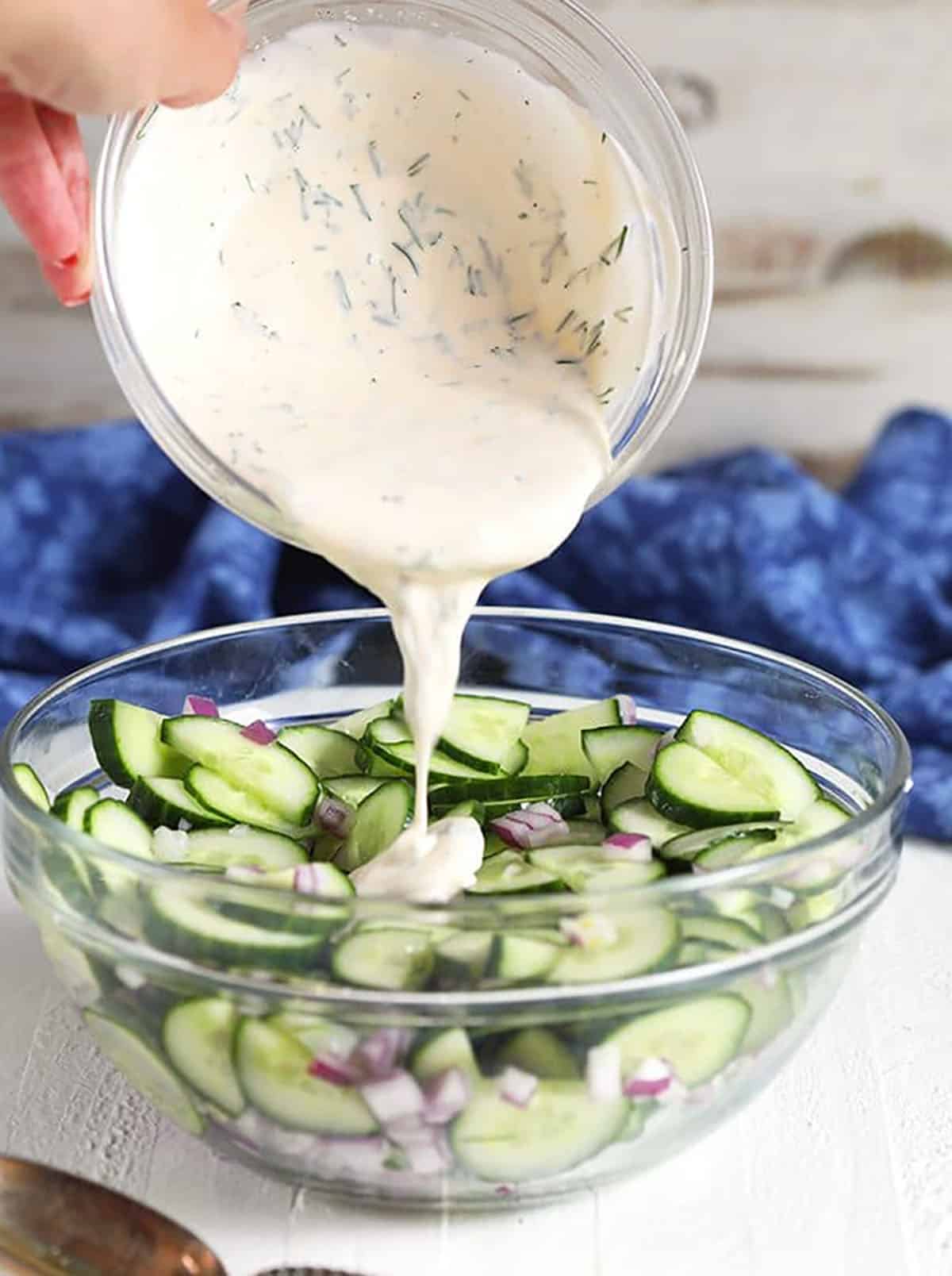 Creamy Cucumber Salad Dressing being poured over cucumbers in a glass bowl.