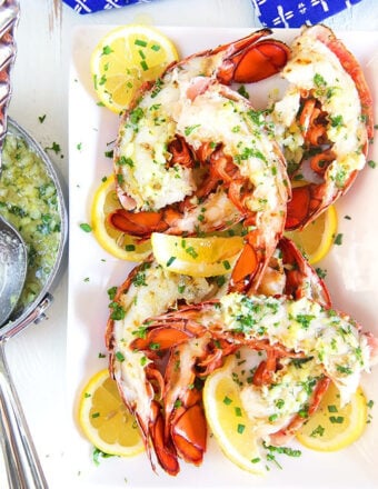 grilled lobster tails on a white platter with a pan of garlic butter sauce next to it.