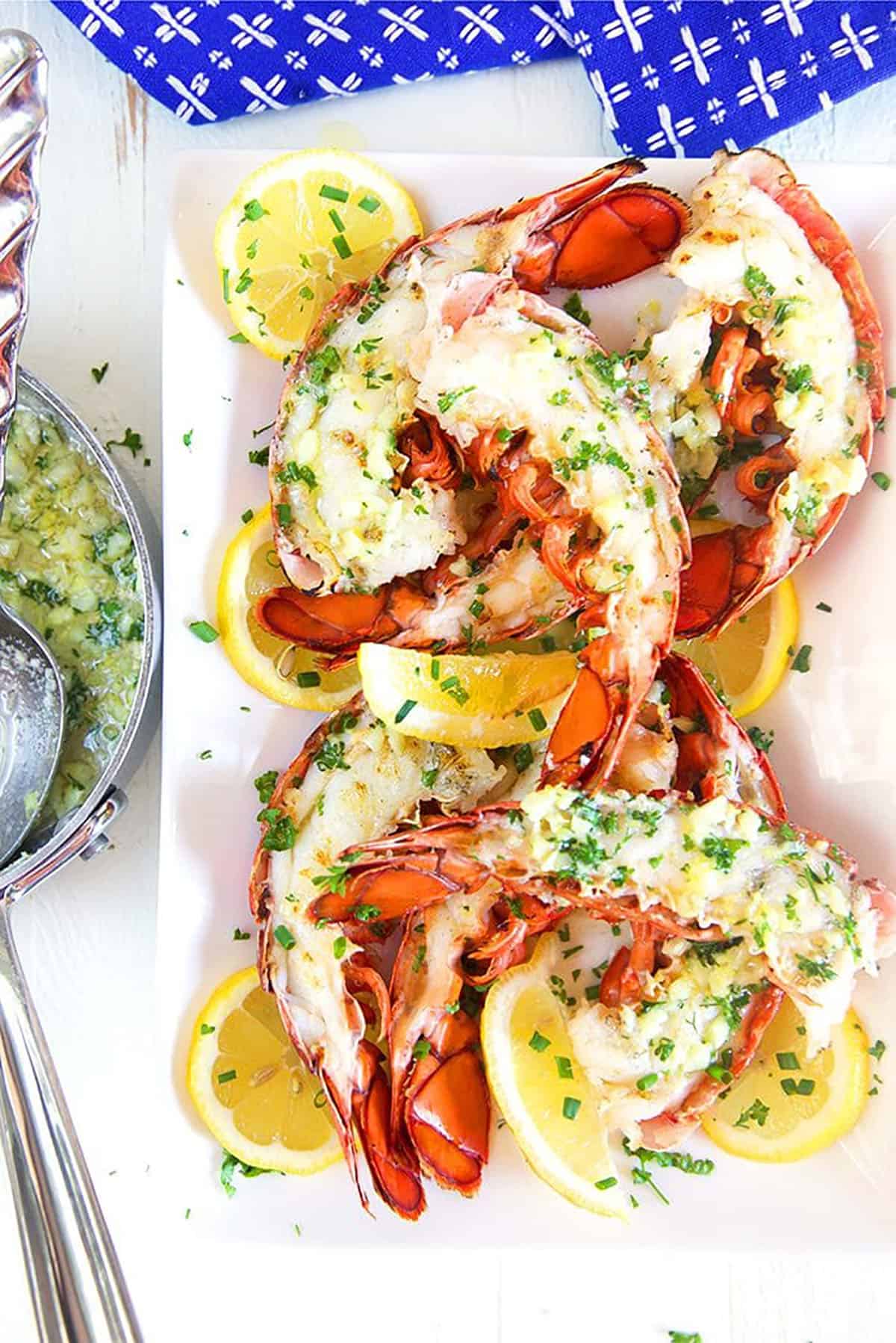 grilled lobster tails on a white platter with a pan of garlic butter sauce next to it.