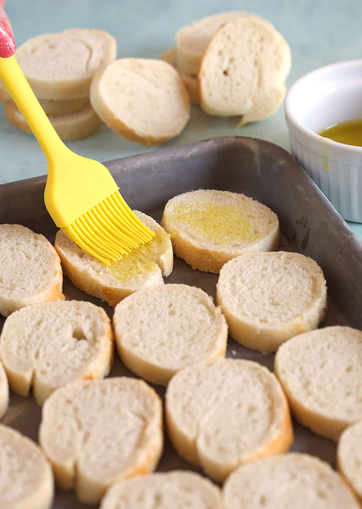 A yellow rubber brush is brushing butter onto slice of crostini. 