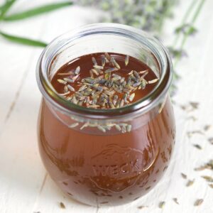 A small glass jar of simple syrup is placed on a white surface with lavender surrounding it.
