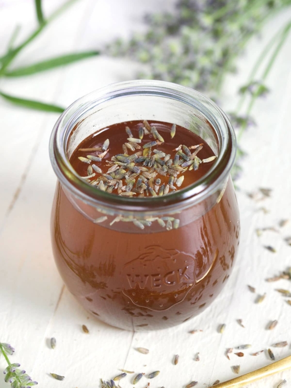 A small glass jar of simple syrup is placed on a white surface with lavender surrounding it.