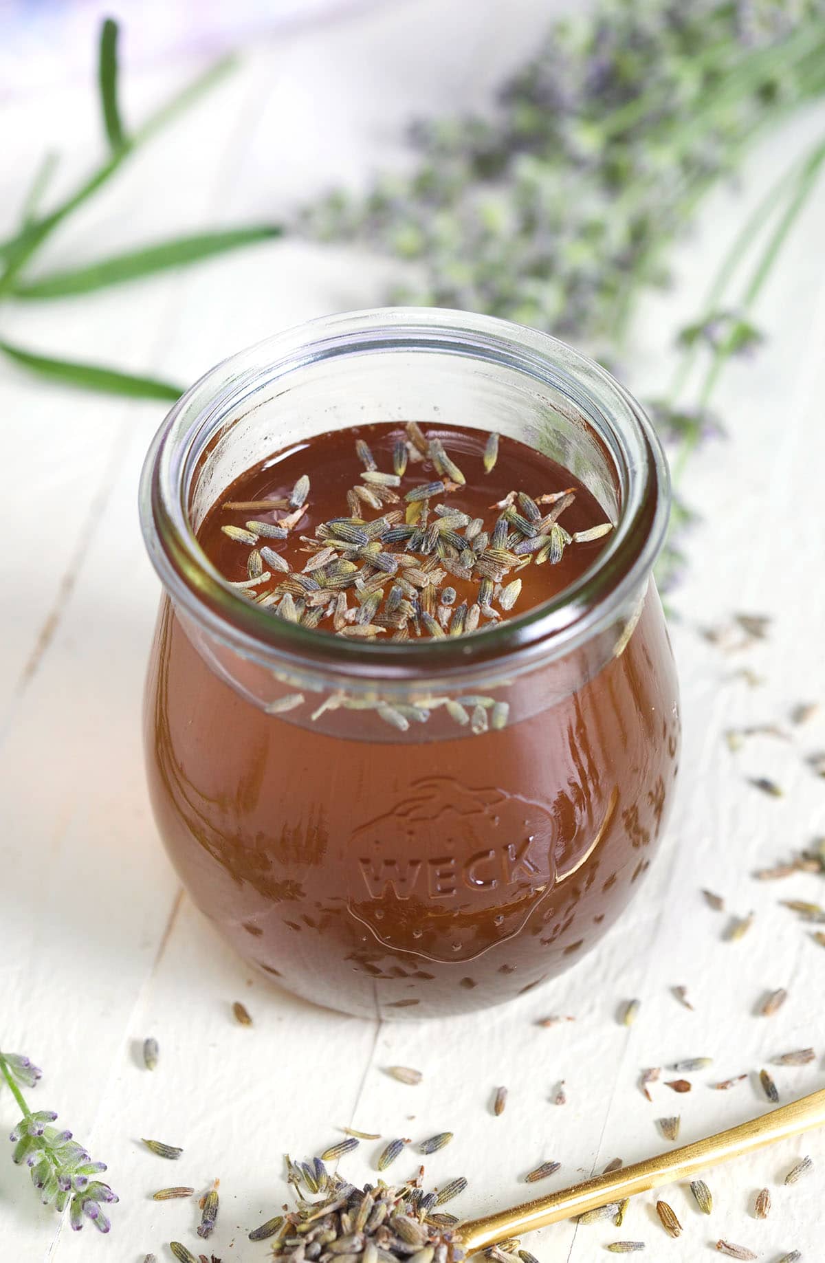 A small glass jar of simple syrup is placed on a white surface with lavender surrounding it.